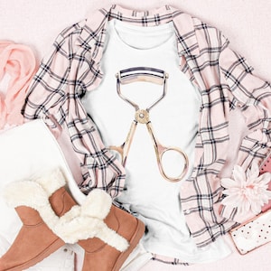 May include: A white t-shirt with a gold eyelash curler graphic is displayed with a pink plaid shirt, brown boots with faux fur trim, a pink scarf, and a pink notebook. The items are arranged on a pink surface.