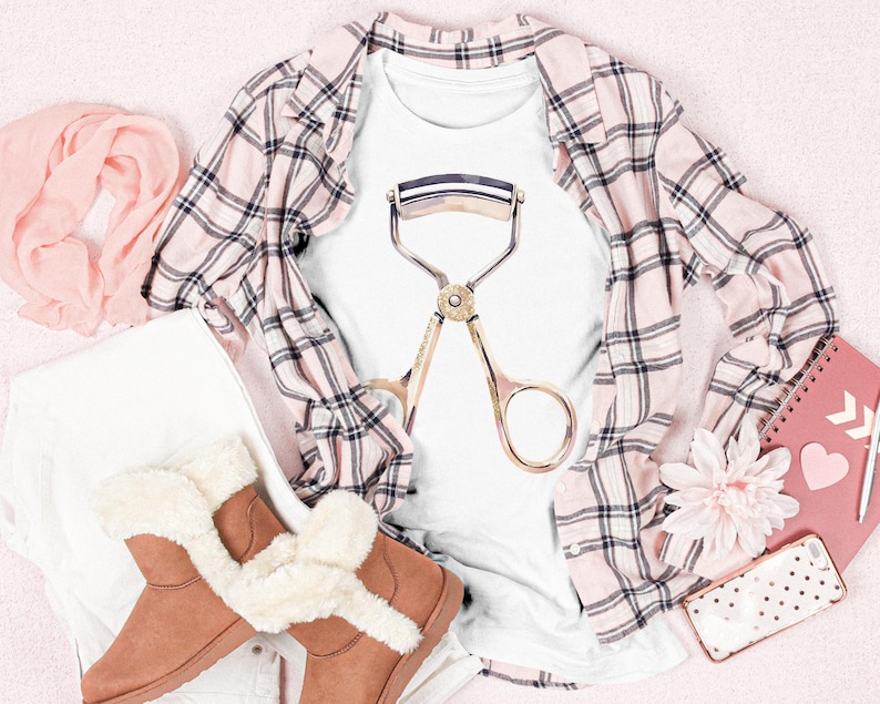 May include: A white t-shirt with a gold eyelash curler graphic is displayed with a pink plaid shirt, brown boots with faux fur trim, a pink scarf, and a pink notebook. The items are arranged on a pink surface.