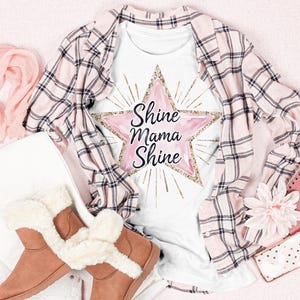 K&ouml;nnte beinhalten: Draufsicht mit einem wei&szlig;en T-Shirt mit einem rosa Sterndesign und dem Text "Shine Mama Shine". Das Shirt wird mit einem rosa karierten Hemd, einer wei&szlig;en Hose, braunen Stiefeln, einem rosa Schal, einem rosa Notizbuch und einer Handyh&uuml;lle kombiniert.