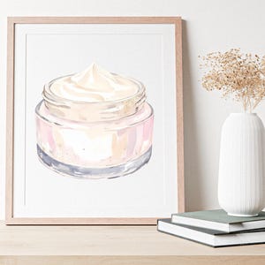 May include: Watercolor illustration of a jar of cream with a light pink and gold design. The jar is displayed in a wooden frame, with a white vase and books in the background.