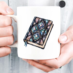 May include: White ceramic mug featuring a detailed illustration of a closed journal. The journal has a dark cover with a geometric design in shades of blue, teal, and purple, with glitter accents. A blue ribbon bookmark is visible.