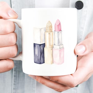 May include: White ceramic mug featuring a watercolor illustration of three lipsticks. The lipsticks are in shades of navy, gold glitter, and pink. The mug is held by a person wearing a light blue shirt.