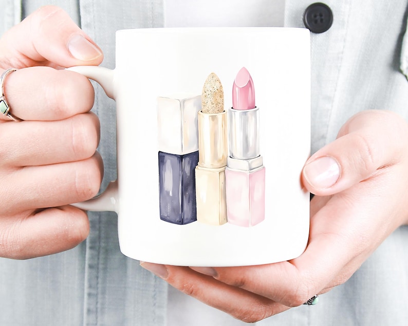 May include: White ceramic mug featuring a watercolor illustration of three lipsticks. The lipsticks are in shades of navy, gold glitter, and pink. The mug is held by a person wearing a light blue shirt.