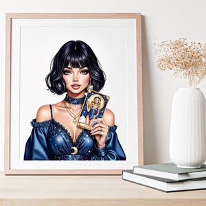 Pode incluir: Impress&atilde;o art&iacute;stica emoldurada com uma mulher de cabelo escuro e um vestido azul segurando uma carta de tar&ocirc;. A obra de arte tem um tema celestial com estrelas e uma lua crescente. A impress&atilde;o &eacute; exibida em uma mesa de madeira com um vaso branco e livros.