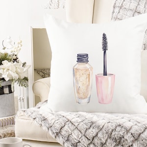 May include: White decorative pillow featuring a watercolor illustration of makeup. The design includes a bottle of foundation and a mascara wand with a pink container. The pillow is on a white couch with a gray, textured blanket.