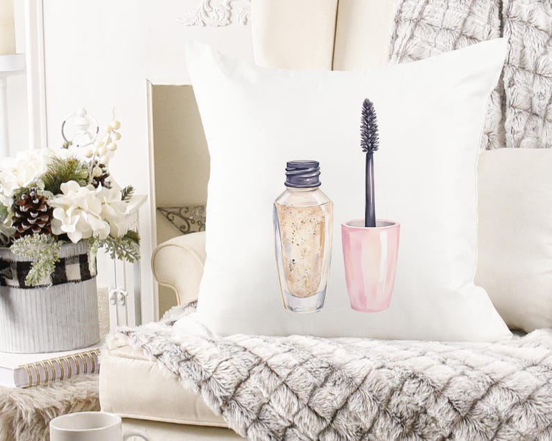 May include: White decorative pillow featuring a watercolor illustration of makeup. The design includes a bottle of foundation and a mascara wand with a pink container. The pillow is on a white couch with a gray, textured blanket.