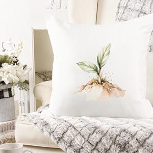 May include: White square throw pillow featuring a watercolor illustration of a small plant with green and white leaves. The plant is emerging from a mound of earth with gold glitter accents. The pillow is on a white couch with a gray blanket.