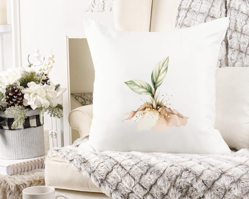 May include: White square throw pillow featuring a watercolor illustration of a small plant with green and white leaves. The plant is emerging from a mound of earth with gold glitter accents. The pillow is on a white couch with a gray blanket.