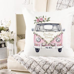 K&ouml;nnte beinhalten: Quadratisches wei&szlig;es Kissen mit einer rosa-wei&szlig;en Vintage-Van-Illustration. Der Van ist mit floralen Akzenten und dem Text "Roadtrip Mama" verziert. Das Nummernschild lautet "MAMA".