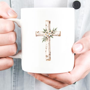 May include: White ceramic mug featuring a cross design. The cross is made to look like birch wood and is decorated with white flowers and green leaves. The mug is held by a person.