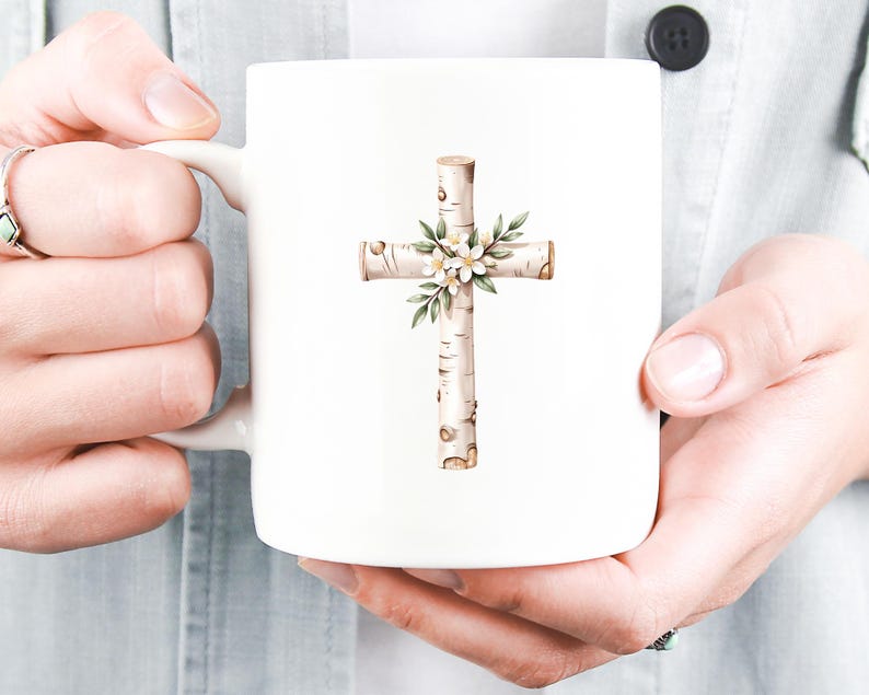 May include: White ceramic mug featuring a cross design. The cross is made to look like birch wood and is decorated with white flowers and green leaves. The mug is held by a person.