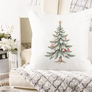 Puede incluir: Coj&iacute;n cuadrado blanco con un dise&ntilde;o de &aacute;rbol de Navidad en acuarela. El &aacute;rbol est&aacute; decorado con adornos rosas y blancos y coronado con una estrella dorada. El coj&iacute;n est&aacute; sobre una manta afelpada con estampado gris y blanco.