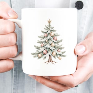 Puede incluir: Taza de cer&aacute;mica blanca con una ilustraci&oacute;n de acuarela de un &aacute;rbol de Navidad adornado con adornos rosas y dorados y una estrella dorada. El &aacute;rbol tiene agujas verdes y un tronco marr&oacute;n. La taza est&aacute; sostenida por una persona.