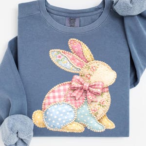 May include: A blue Comfort Colors sweatshirt featuring a patchwork bunny design. The bunny has pink, blue, and yellow patches with floral and polka dot patterns, and a pink gingham bow. A matching blue scrunchie rests on the shoulder.