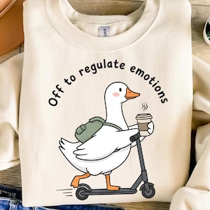 May include: Cream-colored sweatshirt featuring a cartoon goose riding a scooter and holding a coffee cup. The goose wears a backpack and the text "Off to regulate emotions" arches above the goose.
