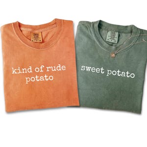 May include: Two crew neck t-shirts are shown, one orange and one green. The orange shirt has the text "kind of rude potato" in white. The green shirt has the text "sweet potato" in white. A necklace is on the green shirt.