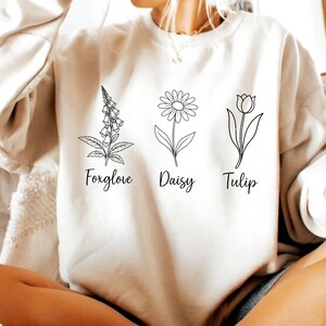 May include: White sweatshirt with a graphic of three flowers: a foxglove, a daisy, and a tulip. The flowers are outlined in black and the names of the flowers are written below each flower.