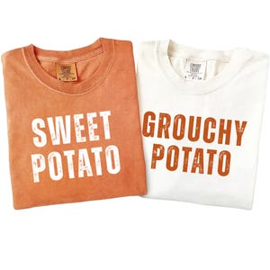 May include: Two crew neck t-shirts. One is a burnt orange color with white text reading SWEET POTATO. The other is white with burnt orange text reading GROUCHY POTATO. Both shirts feature Comfort Colors brand labels at the inner neckline.
