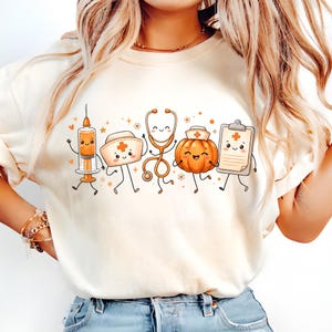 May include: A cream-colored t-shirt featuring a graphic print of five anthropomorphic medical items: a syringe, a nurse cap, a stethoscope, a pumpkin wearing a nurse cap, and a clipboard, all decorated with small orange hearts and stars.