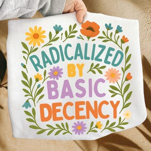 Könnte beinhalten: Weißes rechteckiges Handtuch mit dem Text "RADICALIZED BY BASIC DECENCY" in Türkis, Lila, Orange und Gelb. Der Text ist von einem floralen Kranz aus gelben, orangen, blauen und lila Blumen mit grünen Blättern umgeben.
