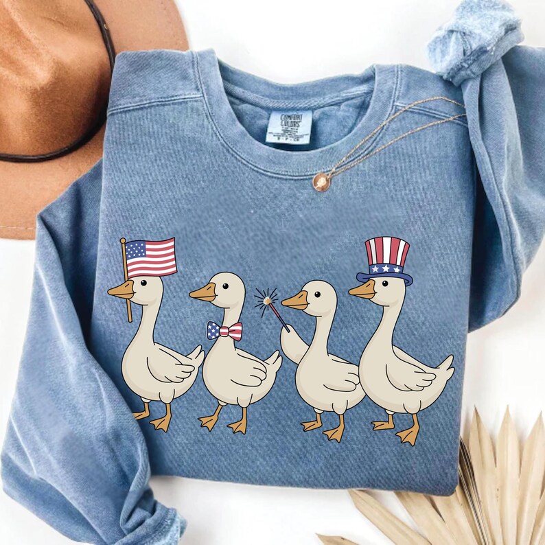 May include: A blue sweatshirt featuring a graphic of four cartoon ducks. Each duck is decorated with patriotic accessories, including an American flag, bow tie, sparkler, and top hat. The sweatshirt is a solid blue color.
