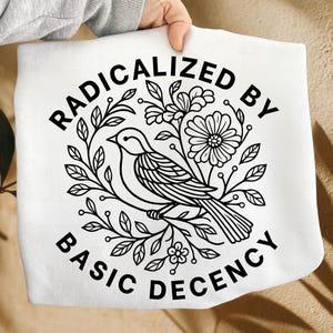May include: White tote bag with a black and white bird and floral design. The text "RADICALIZED BY BASIC DECENCY" is printed in a circular pattern around the bird and flowers.