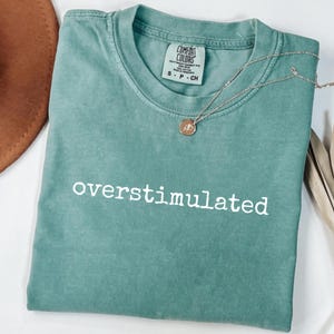 May include: A teal Comfort Colors t-shirt with the word "overstimulated" printed in white. A gold-colored necklace with a pendant is draped across the shirt. A brown hat is partially visible in the upper left corner.