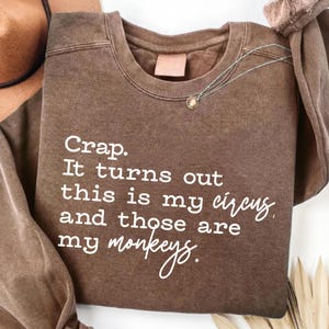 May include: A brown crew neck sweatshirt with the white text: "Crap. It turns out this is my circus and those are my monkeys." A silver necklace with a small pendant is visible. The sweatshirt is folded.