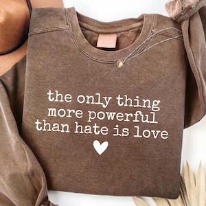 May include: A brown sweatshirt with the text "the only thing more powerful than hate is love" in white, along with a white heart graphic. A necklace with a small pendant is visible. The sweatshirt is part of a clothing collection.