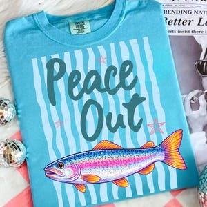 May include: A light blue t-shirt with the words "Peace Out" in teal script, set against a background of vertical blue stripes. A colorful fish illustration with orange and yellow fins is at the bottom. The shirt has a round neckline.