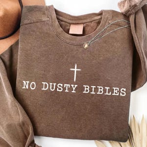 Può includere: Una felpa marrone con la scritta "NO DUSTY BIBLES" e una croce bianca. La felpa girocollo ha una vestibilità ampia ed è abbinata a una collana. La felpa è di colore neutro.