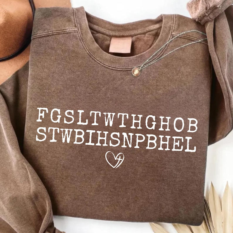 May include: A brown crewneck sweatshirt with white text and a heart symbol. The text reads "FGSLTWTGHOB STWBIHSNPBHEL". A silver necklace with a small pendant is visible. The sweatshirt is a neutral color.