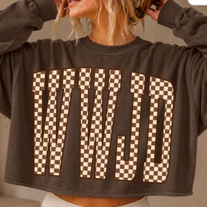 May include: A charcoal gray cropped sweatshirt featuring the letters WWJD in a large, bold font. The letters are filled with a brown and white checkered pattern. The garment has a crew neckline, long sleeves with ribbed cuffs, and a raw hemline.