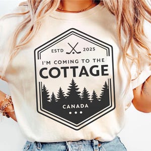 May include: Cream-colored t-shirt featuring a black graphic with the text "I'M COMING TO THE COTTAGE" and "CANADA". The graphic includes crossed hockey sticks, the year 2025, and a silhouette of pine trees. The shirt has a crew neck.