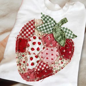 Könnte beinhalten: Weißes T-Shirt mit einem großen Erdbeer-Patchwork-Design. Die Erdbeere besteht aus verschiedenen roten und rosa Stoffen, darunter Vichy, Blumenmuster und Herzen. Grüne Vichy-Blätter krönen die Erdbeere.