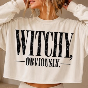 May include: A cream-colored long-sleeve cropped sweatshirt featuring the black distressed-style text "WITCHY, OBVIOUSLY." printed across the chest. The garment has a crew neckline, ribbed cuffs, and a raw-edge hemline at the waist.