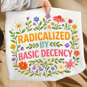 Może przedstawiać: Biała torba typu tote z napisem "RADICALIZED BY BASIC DECENCY" w kolorze pomarańczowym, turkusowym i różowym, otoczona kwiatowym wieńcem z kolorowych kwiatów i zielonych liści. Torba jest trzymana przez osobę.