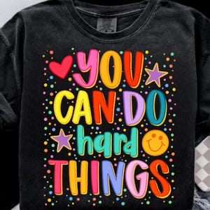 May include: Black t-shirt with the phrase "YOU CAN DO HARD THINGS" in colorful, bubble letters. The design includes a red heart, stars, and a smiling face, all surrounded by colorful dots.