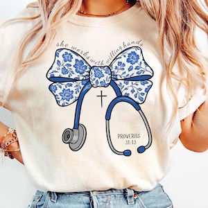 May include: Cream-colored t-shirt featuring a blue floral bow and a stethoscope design. The design includes a cross and the text "PROVERBS 31:13". The words "she works with willing hands" are arched above the bow.