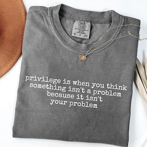May include: A gray Comfort Colors t-shirt with the text "privilege is when you think something isn't a problem because it isn't your problem" in white. A silver necklace with a pendant is on the shirt.