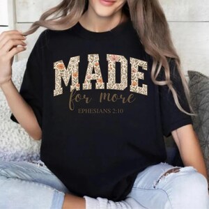 Op de afbeelding: Zwart T-shirt met een bloemenprint met de tekst "MADE for more" en "EPHESIANS 2:10" in bruine letters.