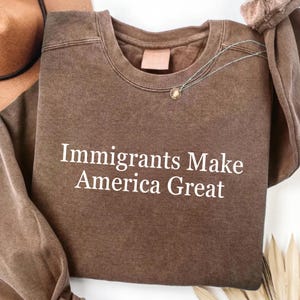 Könnte beinhalten: Ein braunes Sweatshirt mit dem weißen Text "Immigrants Make America Great". Das Sweatshirt mit Rundhalsausschnitt hat lange Ärmel. Eine Halskette mit einem kleinen Anhänger ist auf dem Shirt drapiert.