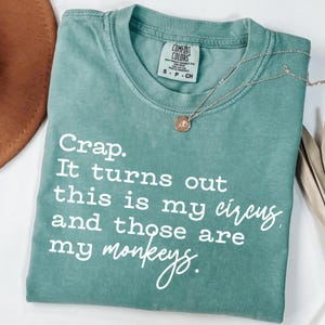 以下が含まれることがあります： 「Crap. It turns out this is my circus and those are my monkeys」の白文字がプリントされた、シーフォームグリーンのComfort Colors Tシャツ。小さなペンダントが付いたシルバーのネックレスがシャツの上にあります。
