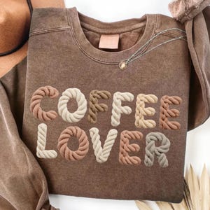 May include: A brown sweatshirt with the words "COFFEE LOVER" in a 3D rope-like font. The letters are in various shades of brown, beige, and cream. A silver necklace with a small pendant is visible near the neckline. The sweatshirt is folded.