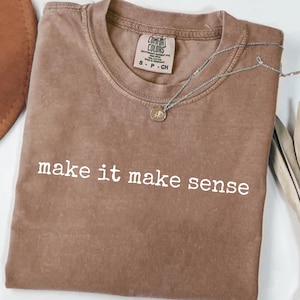 以下が含まれることがあります： 「make it make sense」という白い文字がプリントされた茶色のTシャツ。ペンダント付きのシルバーのネックレスがシャツの上にあります。シャツにはComfort Colorsのタグが付いています。