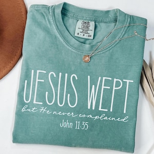 May include: A seafoam green t-shirt with the text "JESUS WEPT" in large white letters. Below, in smaller white script, it reads "but He never complained" and "John 11:35". A silver necklace with a pendant is draped across the shirt.