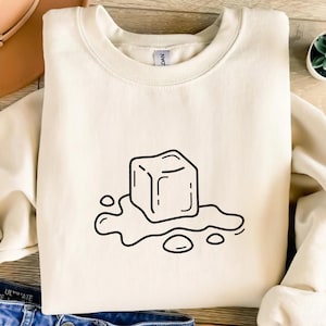 Puede incluir: Sudadera de color crema con un dibujo lineal negro de un cubo de hielo derritiéndose. El cubo de hielo tiene forma cuadrada con bordes redondeados, y el agua derretida se representa alrededor. La sudadera tiene cuello redondo y mangas largas.