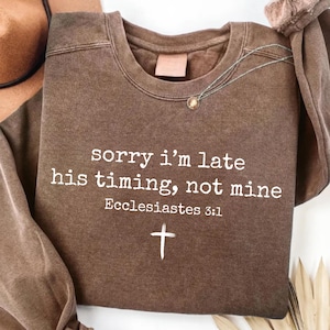 May include: A brown crewneck sweatshirt with the text "sorry i'm late his timing, not mine" and the Bible verse Ecclesiastes 3:1, along with a cross symbol. A necklace with a small pendant is visible.