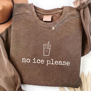 以下が含まれることがあります： 「no ice please」の白い文字と、カップの白い線画がプリントされた茶色のスウェットシャツ。小さなペンダントが付いたネックレスがスウェットシャツにかけられています。
