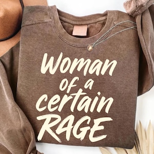 May include: A brown sweatshirt with the text "Woman of a certain RAGE" in a cream-colored font. The sweatshirt has a crew neck and long sleeves. A silver necklace with a small pendant is draped across the front.